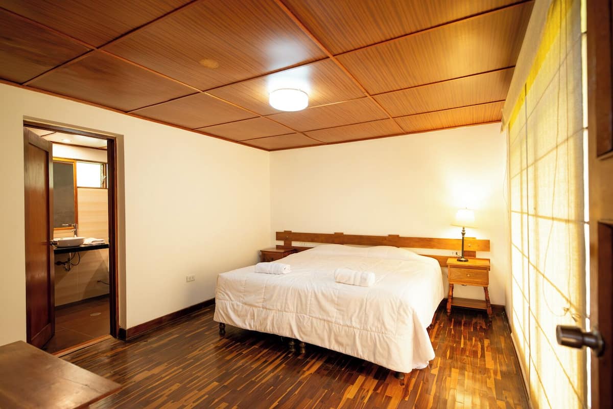 A hotel room with a wooden ceiling, a bed with a white bedspread, a bedside table with a lamp, and a wooden floor.