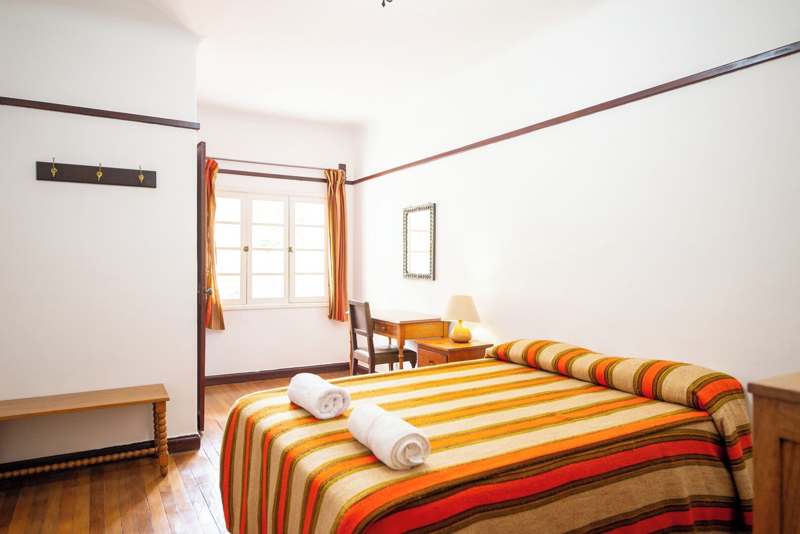 A hotel room with a bed, a desk, a chair, a window with curtains, and a bench.