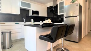 A modern kitchen with white cabinets, a black countertop, a stainless steel refrigerator, and a bar stool. - Sea Turtle Condo 2 Bdr-2 Bth in Palm Beach Area, walking distance to the beach. (Noord)