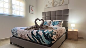 A bedroom with a bed, a nightstand, a lamp, a window, and a wall art piece. - Sea Turtle Condo 2 Bdr-2 Bth in Palm Beach Area, walking distance to the beach. (Noord)