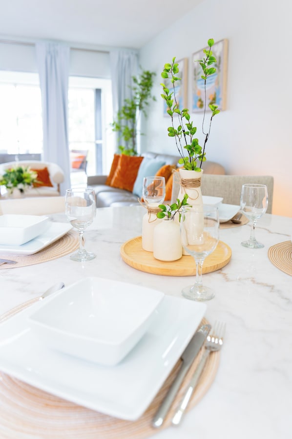 A dining table set with white plates, cutlery, and glasses, with a vase of green plants and candles on a wooden board.