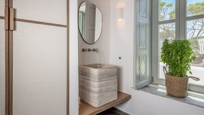 A bathroom with a stone sink, a mirror, a potted plant, and a window with open shutters. - Sunset Plaka (Milos)