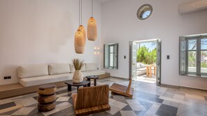 A modern living room with a sofa, a coffee table, and a wooden bench. There are two hanging lights and a window with a view of greenery. - Sunset Plaka (Milos)