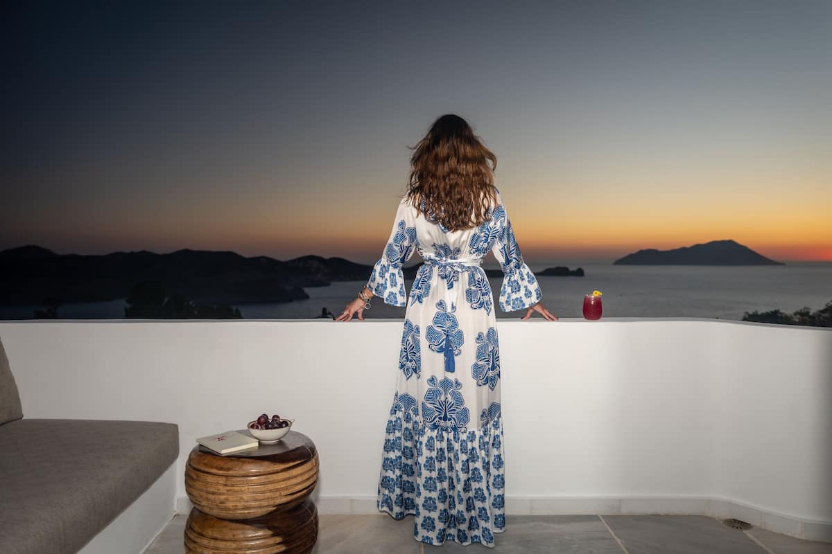 A woman in a patterned dress standing on a balcony overlooking a sunset with mountains in the distance.
