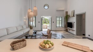 A modern living room with a sofa, a dining table, and a TV mounted on the wall. There is a large window providing natural light and a view of the outdoors. - Sunset Plaka (Milos)