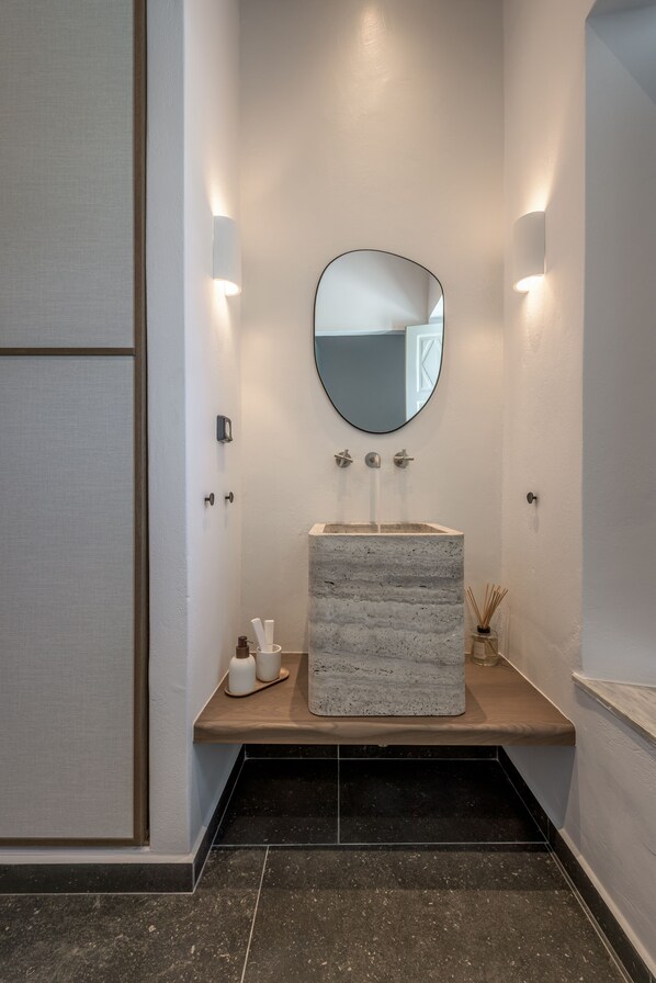 A modern bathroom with a rectangular stone sink, a round mirror, and two wall-mounted lights. - Sunset Plaka (Milos)