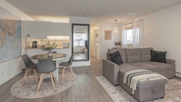 A modern living room with a grey sofa, a dining table with chairs, and a kitchen visible through an open door.