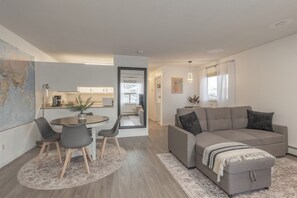 Una sala de estar moderna con un sofá gris, una mesa de comedor con sillas, y una cocina visible a través de una puerta abierta.
