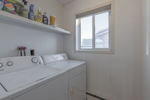 Uma lavanderia com duas máquinas de lavar e secar brancas, uma janela e prateleiras com produtos de limpeza.