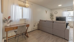 A modern living room with a sofa, a desk with a chair, a television, and a wall-mounted clock.