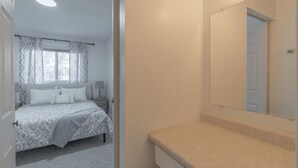 A bedroom with a bed, a bedside table, a nightstand, a window with curtains, and a bathroom with a sink and mirror.