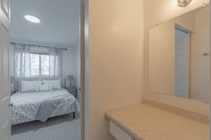 Quarto com cama, mesa de cabeceira, cortinas na janela e banheiro com pia e espelho.