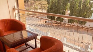 A balcony with a glass table and two orange chairs, overlooking a street with trees and a white fence. - E-Gold Luxury Hotel (Abuja)