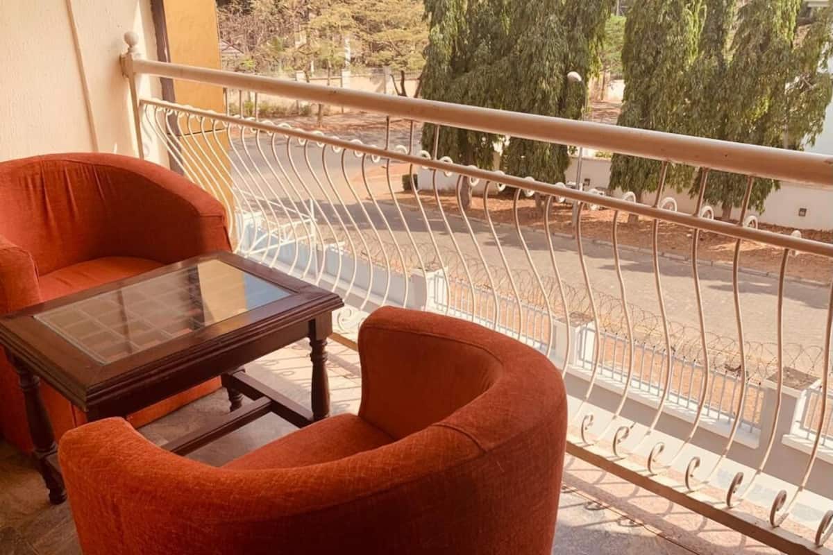 Un balcon avec une table en verre et deux chaises orange, donnant sur une rue bordée d’arbres et délimitée par une clôture blanche.