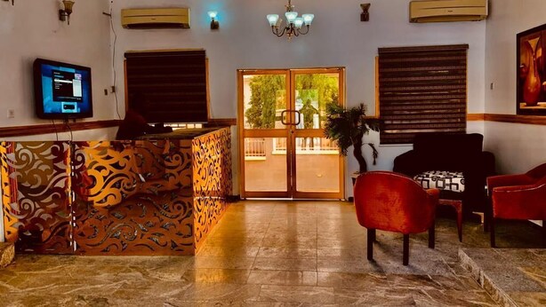 A lobby with a patterned reception desk, a television, a chandelier, and a view through the glass doors. - E-Gold Luxury Hotel (Abuja)
