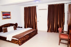 A bedroom with a wooden bed, brown curtains, a desk with a chair, and a painting on the wall. - E-Gold Luxury Hotel (Abuja)