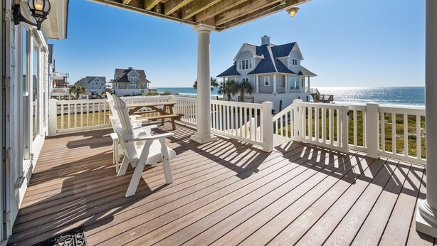 A wooden deck with white railings, a bench, and a view of a large house by the ocean.
