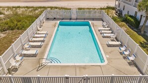 A rectangular swimming pool with a white deck, surrounded by lounge chairs and a wooden fence. A beach and ocean are visible in the background.