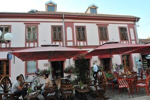 An outdoor café with wooden tables and chairs, red umbrellas, and a building with a tiled roof and shutters.