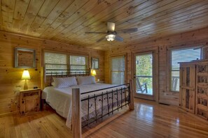 A wooden cabin bedroom with a bed, two bedside lamps, a ceiling fan, and a window with blinds.