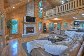 A log cabin interior with a stone fireplace, leather sofas, a ceiling fan, and a balcony.