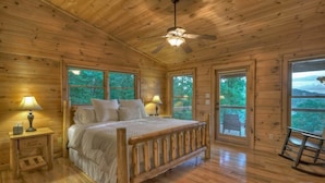 Une chambre dans une cabane en bois, avec un lit, deux tables de chevet, un ventilateur de plafond et de grandes fenêtres.