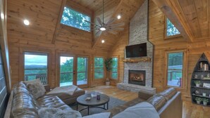 L’intérieur d’un chalet en rondins, avec une cheminée en pierre, des canapés en cuir, une table basse ronde et de grandes fenêtres donnant sur le paysage extérieur.