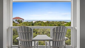Ein Balkon mit zwei Adirondack-Stühlen und einem Tisch, der auf eine Küstenlandschaft mit einem Gebäude und Vegetation blickt.