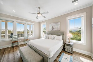A bedroom with a large bed, a ceiling fan, and a view of the outdoors through multiple windows.