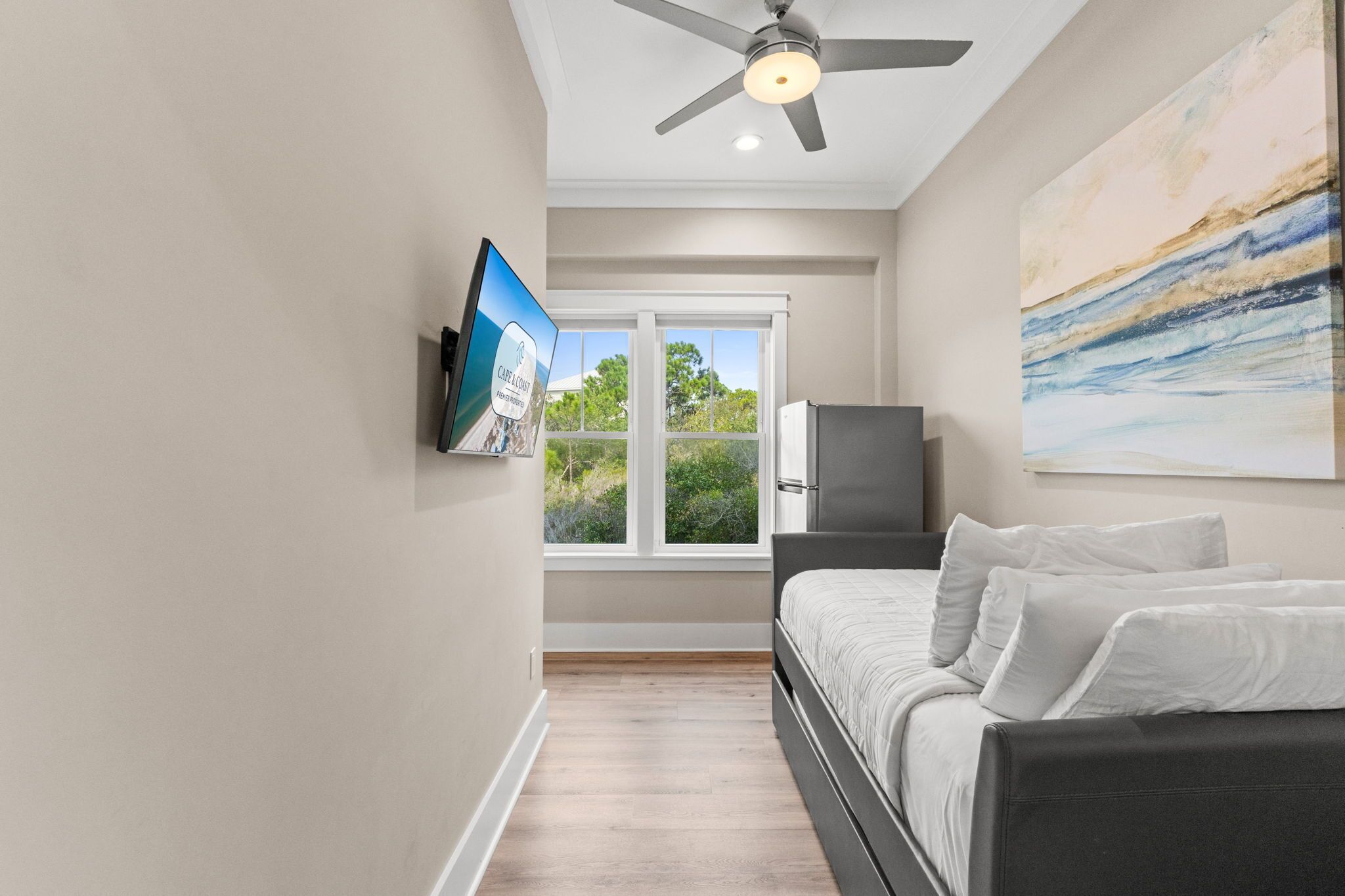 A bedroom with a bed, a television mounted on the wall, a ceiling fan, and a window with a view of greenery.