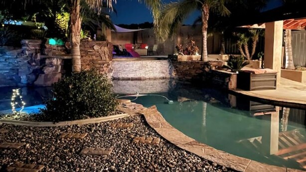 A swimming pool with a waterfall feature, surrounded by palm trees and a stone wall. - SERENITY AWAITS HILL WINE Co#2 (Temecula)