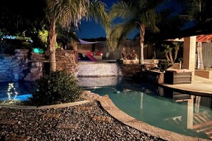 A swimming pool with a waterfall feature, surrounded by palm trees and a stone wall.
