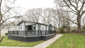 A single-story mobile home with a deck, surrounded by a grassy area and trees. - Owls Nest (Wimborne)