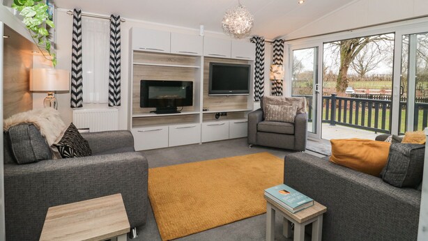 A modern living room with grey sofas, a wooden coffee table, a TV on a white cabinet, a hanging chandelier, and a view of the outdoors through large sliding doors. - Owls Nest (Wimborne)