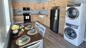 A modern kitchen with wooden cabinets, a stainless steel refrigerator, a double-door washer, and a dining area with a table set for two. - Dune View at Silver Lake (Mears)