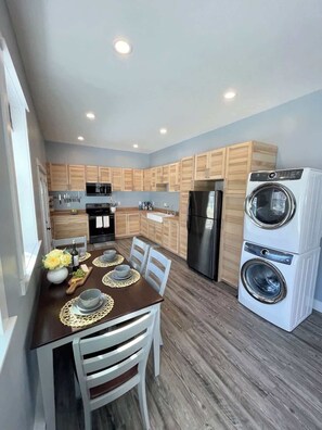 A modern kitchen with wooden cabinets, a stainless steel refrigerator, a double-door washer, and a dining area with a table set for two.