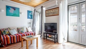 A living room with a wooden floor, a colorful sofa, a TV on a stand, and a painting on the wall.