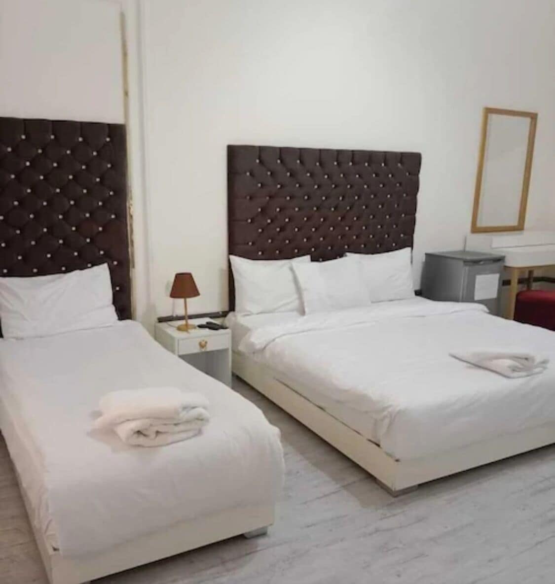 Quarto de hotel com duas camas de solteiro, cada uma com cabeceira e roupa de cama branca. Há uma mesa de cabeceira com abajur e uma pequena mesa com espelho.