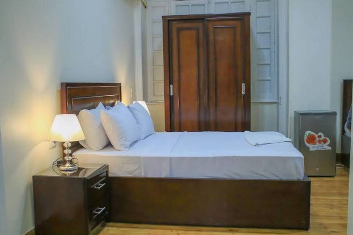 A bedroom with a bed, a nightstand, a lamp, a wardrobe, and a refrigerator.