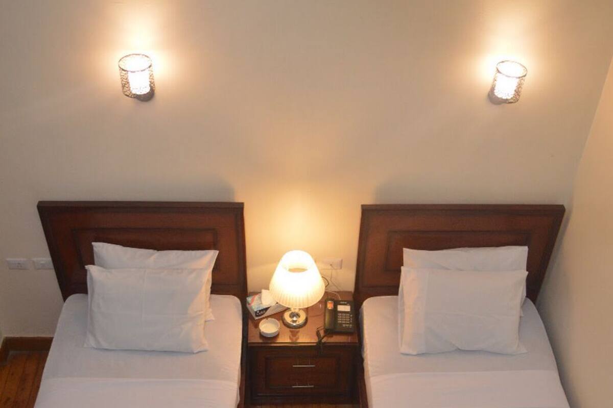 A hotel room with two single beds, each with a wooden headboard, white linens, and towels folded on the beds. There are bedside lamps and a nightstand with a phone and a small table between the beds.