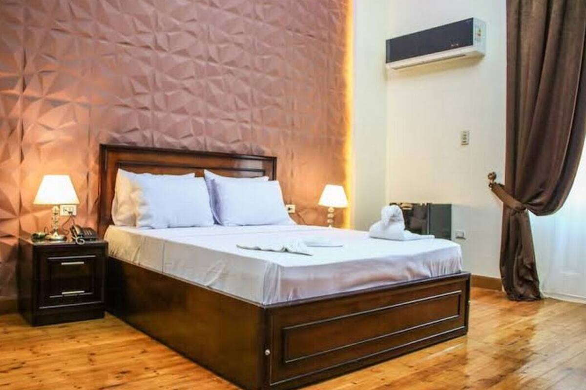 A neatly made bed with a wooden headboard, two bedside lamps, a small bedside table with a phone, and a wall-mounted air conditioner.