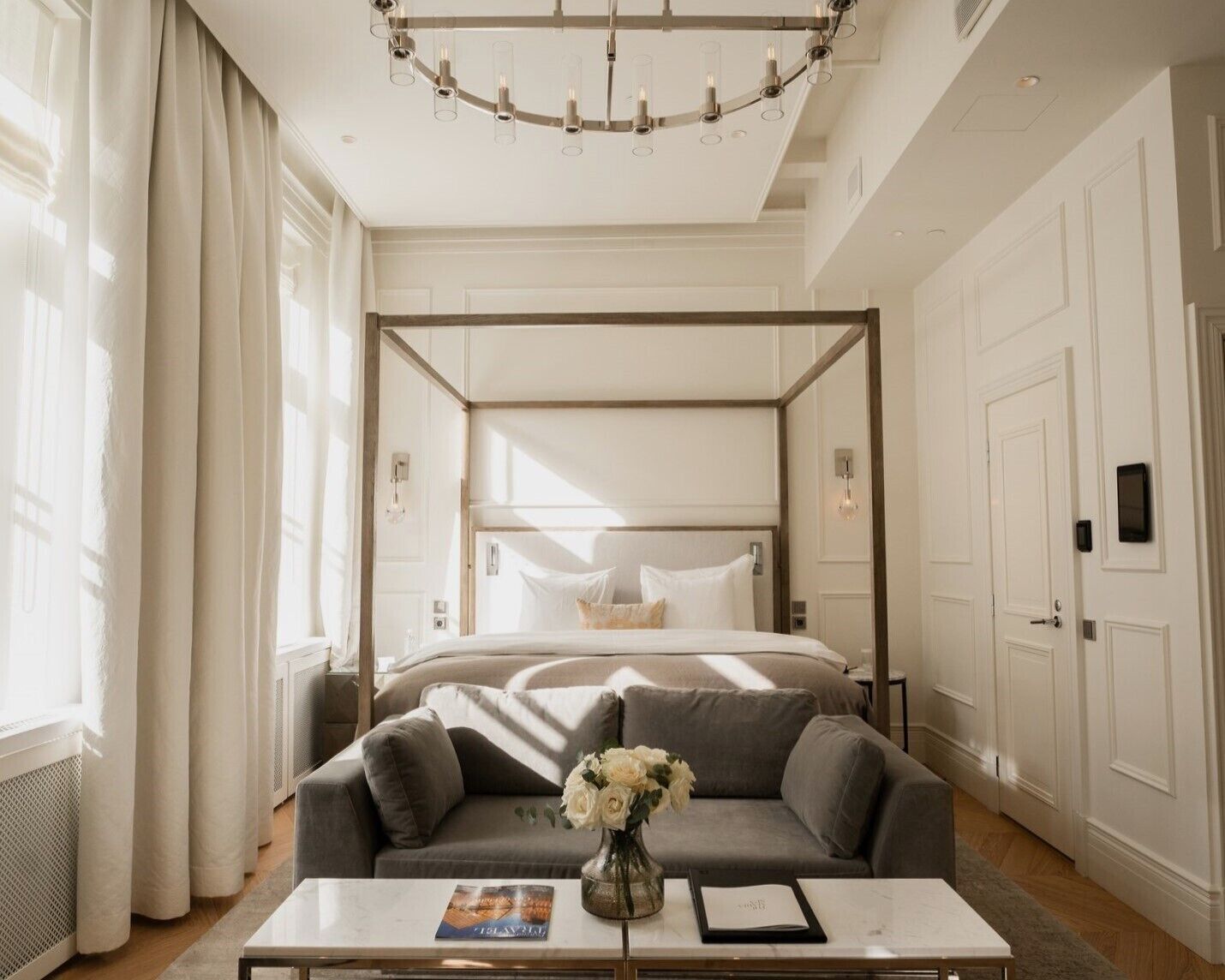 A bedroom with a four-poster bed, a grey sofa, a coffee table with a vase of flowers, and a chandelier.