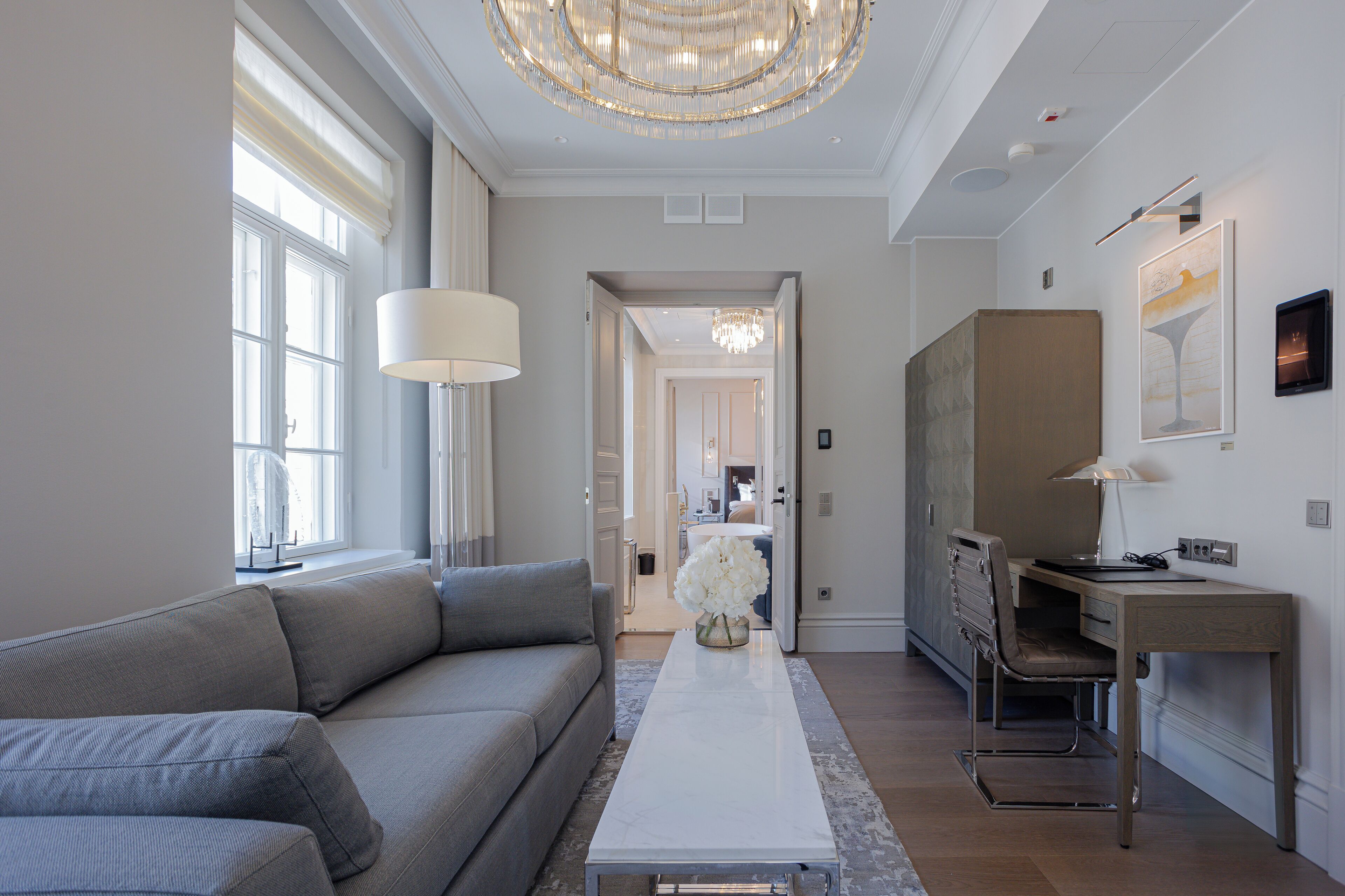 Un salon moderne avec un canapé gris, une table basse blanche et un bureau avec une chaise. Un lustre est suspendu au plafond et un tableau orne le mur.