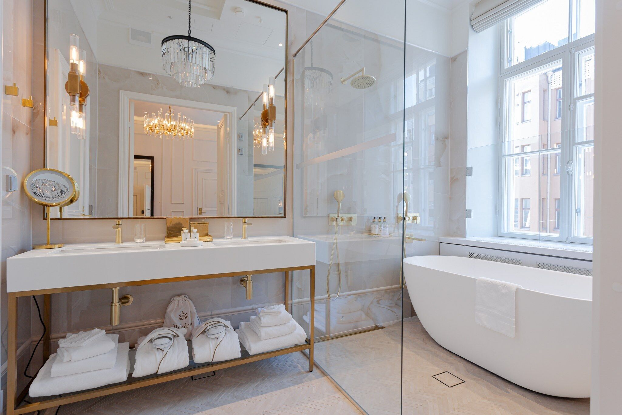 Ein modernes Badezimmer mit einem großen Spiegel, einer goldenen Waschtischkommode, einer freistehenden Badewanne und einem Chandelier.