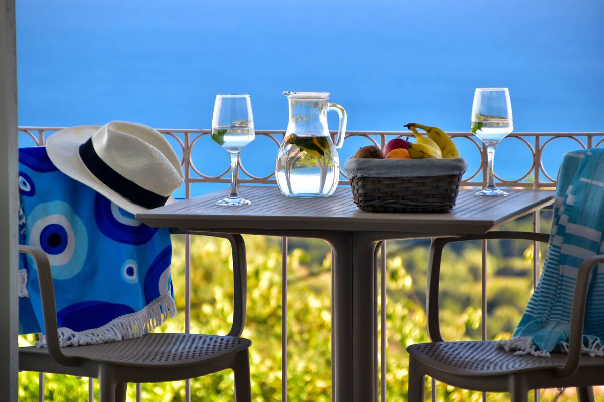 A table with a pitcher, glasses, a hat, and a fruit basket on a balcony with a view.