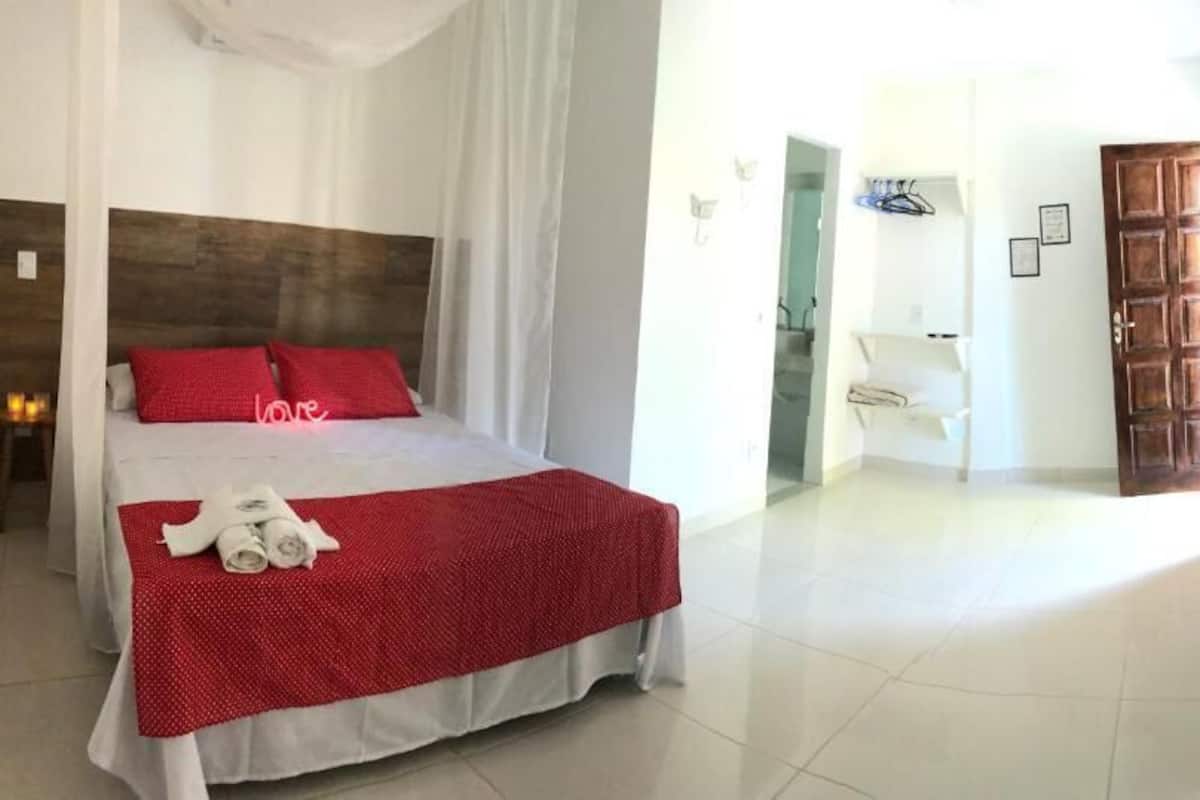Ett hotellrum med en stor säng, en träfärgad sänggavel, ett rött överkast och en vit gardin. I bakgrunden syns en vit stol och en trädörr.