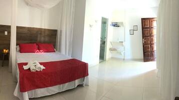A hotel room with a large bed, a wooden headboard, a red bedspread, and a white curtain. There is a white chair and a wooden door in the background.