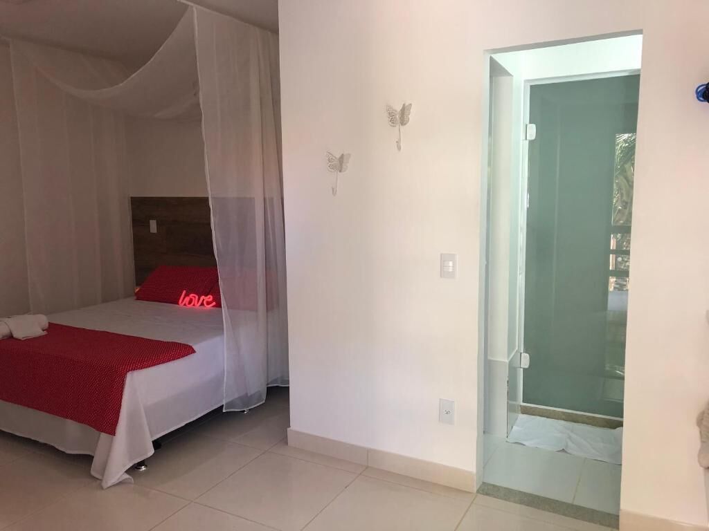 Une chambre à coucher avec un lit recouvert d’un baldaquin blanc, un oreiller rouge orné du mot « amour », et une porte en verre donnant sur une salle de bain.