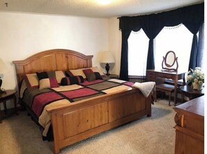 A wooden bed with a headboard, a bedside table, a lamp, a dresser, a mirror, and a window with curtains.