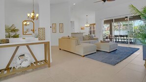 A modern living room with a beige sofa, a white coffee table, and a blue rug. There is a white console table with a mirror and decorative items. A ceiling fan is visible, and the room has large windows.
